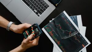 Hands using smartphone beside laptop with stock charts, showcasing digital trading.